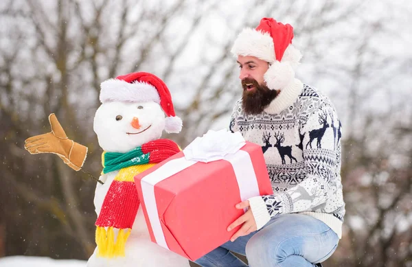 Hipster with beard hold gift box. Surprise concept. Winter games. Winter activity. Winter vacation. Man made snowman. Man Santa hat having fun outdoors. Guy happy face snowy nature background — Stock Photo, Image