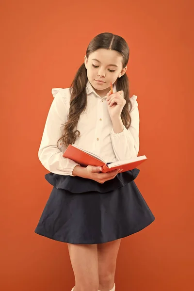 Klein meisje in school uniform. krijg informatieformulier boek. terug naar school. klein kind concentreerde zich op het werk. les lezen. schoolmeisje schrijven notities over oranje achtergrond. voorbereiden voor het lezen van examens — Stockfoto