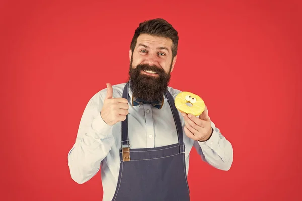 Café et concept de boulangerie. Doux beignet du boulanger. Homme barbu boulanger dans tablier de cuisine tenir dessert mignon. Façons de réduire la faim et l'appétit. Hipster barbu boulanger tenir beignet glacé sur fond rouge — Photo