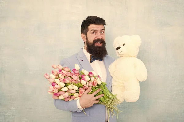 Feliz y enamorada. Caballero haciendo una sorpresa romántica para ella. Un regalo romántico para caballeros. Saludos de cumpleaños. Feliz cumpleaños, mi amor. Hombre barbudo caballero traje pajarita agarre osito de peluche y ramo —  Fotos de Stock