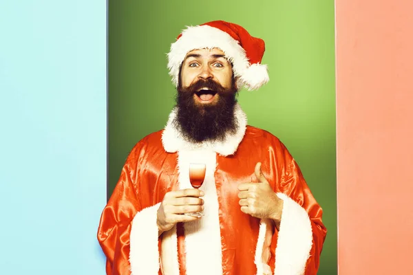 Schöner bärtiger Weihnachtsmann mit langem Bart auf lächelndem Gesicht mit einem Glas alkoholischen Schnaps in rotem Weihnachtsmantel und Neujahrsmütze auf buntem Studiohintergrund — Stockfoto