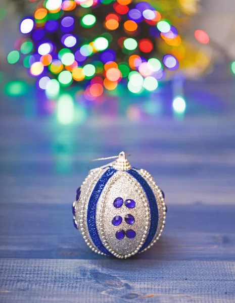 Christmas ornaments concept. Ball ornament on blue wooden surface. Pick colorful decor for christmas tree. Christmas ornament single ball on defocused garland colorful lights background