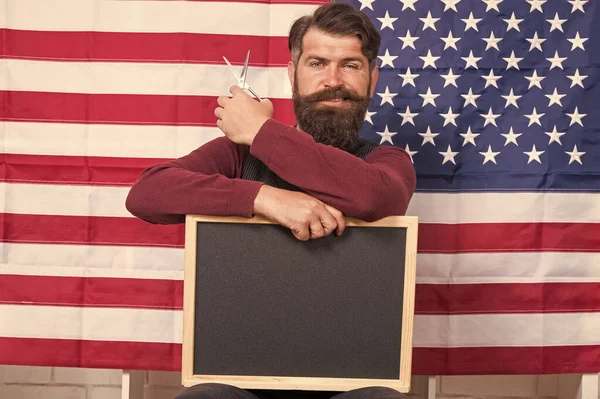 Providing training course. Hipster holding scissors and blackboard in training school. Bearded man giving training course in barbering on usa flag background. Barber school and training