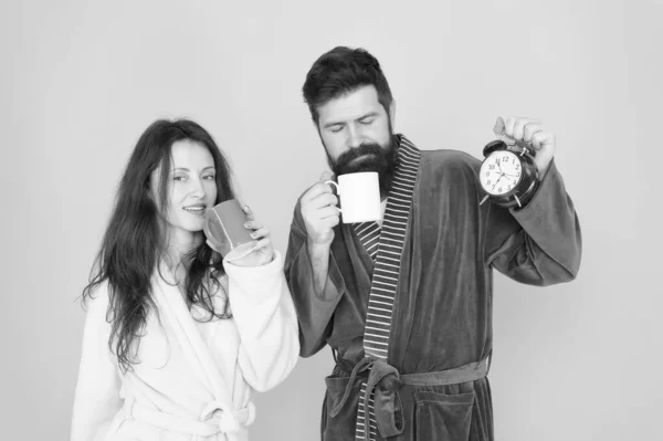 Homme barbu et femme endormie dégustent du café ou du thé le matin. C'est l'heure de se réveiller et de passer une bonne journée. Guy mari tient café et horloge. Chaque matin commence par un café. Couple en peignoirs avec tasses — Photo