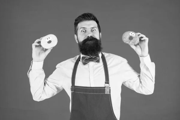 Catering bakery products to you. Bearded man holding ring donuts in bakery shop. Professional baker with glazed bakery food on red background. The perfect doughnuts from our bakery