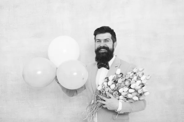 Un rendez-vous romantique. Salutations d'anniversaire. Livraison de fleurs. Pour quelqu'un de spécial. Homme barbu costume gentleman noeud papillon tenir des ballons à air et bouquet. Messieurs faisant une surprise romantique pour elle — Photo