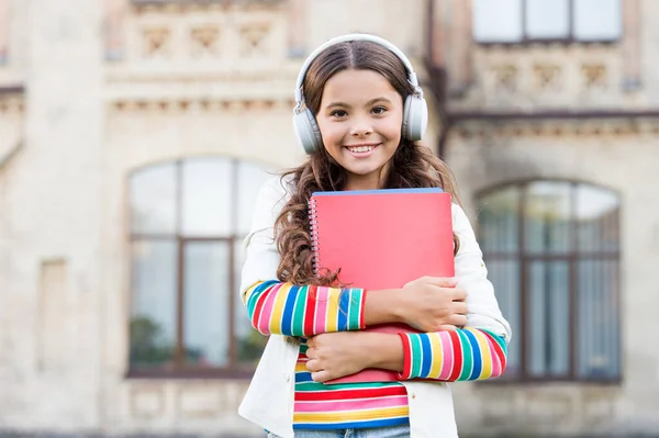 Programas de áudio para alunos. Menina feliz ouvir áudio livro tocando em fones de ouvido. Criança pequena desfrutando de aprendizagem de áudio. Criança em fones de ouvido estéreo segurando materiais de áudio para lição — Fotografia de Stock