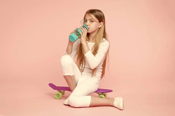 El agua le da energía. Niña patinadora bebiendo agua sobre fondo rosa. Niño sediento bebiendo agua fresca de la botella de plástico. Niña bebiendo de una botella de agua sentada en un penique — Foto de Stock