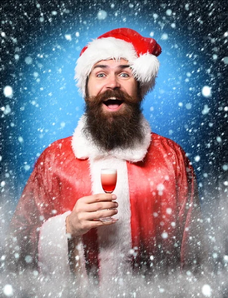 Knappe bebaarde kerstman met lange baard op een glimlachend gezicht met een glas alcoholische shot in rode kerst of kerst trui en nieuwjaarshoed op blauwe studio backgrou — Stockfoto