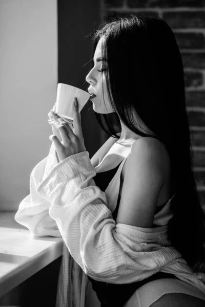 The best coffee for her daily routine. Sensual girl drinking her favorite morning coffee. Pretty woman drinking fresh hot coffee at window. Sexy girl holding ceramic coffee cup