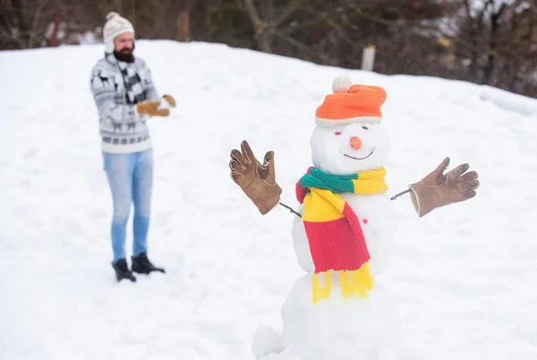 Vacaciones de Navidad. Estilo de vida activo. Juegos de nieve. Ocio al aire libre. Muñeco de nieve y alegre barbudo hipster sombrero de punto y guantes calientes juegan con la nieve al aire libre. Diviértete. Deja que nieve — Foto de Stock