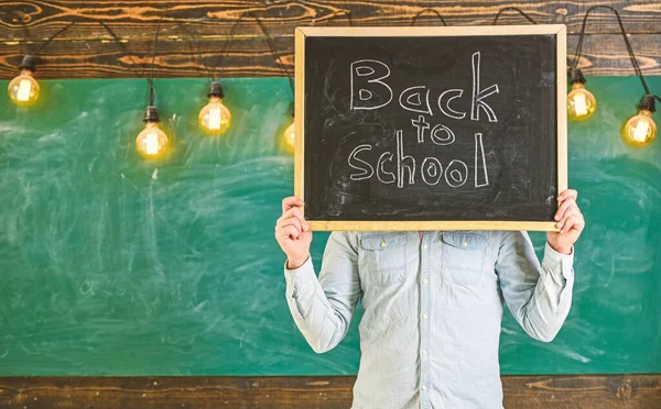Start of school year concept. Teacher holds chalkboard in front of face. Man welcomes students, chalkboard on background. Teacher faceless holds blackboard with title back to schoo