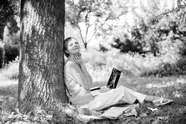 Eğitim teknolojisi ve internet konsepti. Kız parkta dizüstü bilgisayar ile çim üzerinde oturup çalışır. Doğal çevre ofisi. Açık havada çalışmanın faydaları. Dizüstü bilgisayar işi açık havada yalın ağaç ile Kadın. Dinlenmek için dakika