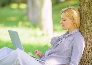 Kız parkta dizüstü bilgisayar ile çim üzerinde oturup çalışır. Doğal çevre ofisi. Açık havada çalışmanın faydaları. Eğitim teknolojisi ve internet konsepti. Dizüstü bilgisayar işi açık havada kadın ağaç gövdesi yalın