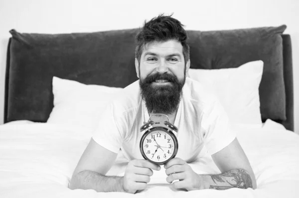 Why you should wake up early every morning. Health benefits of rising early. Waking up early gives more time to prepare and be timely. Hipster bearded man lay in bed with alarm clock. Time to wake up