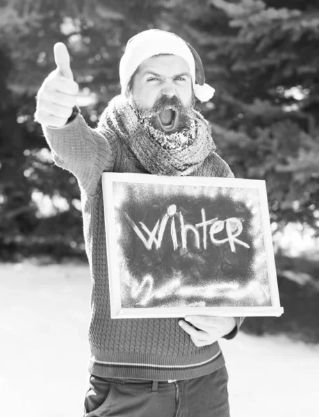 Homem alegre em chapéu de santa claus, hipster barbudo com barba e bigode coberto com geada, dá polegares até gesto com palavra de inverno na placa preta ou quadro no dia ensolarado no backgrou natural — Fotografia de Stock