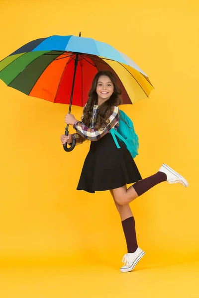 По дорозі до школи. Дощова погода з належним одягом. Весела усміхнена школярка. Дощовий день весело. Щаслива прогулянка під парасолькою. Насолоджуйтесь концепцією дощу. Дитяча дівчинка щаслива тримає барвисту веселку парасольку — стокове фото