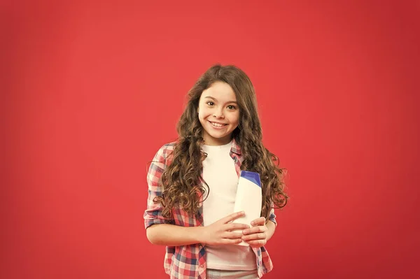 The best shampoo for kids hair. Little girl holding shampoo bottle on red background. Small child with long curly hairstyle choosing shampoo. Shampoo will make your hair grow