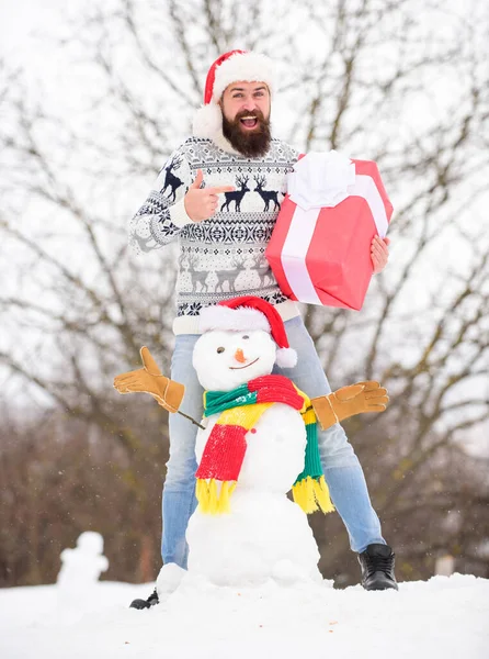 Surprise concept. Winter games. Winter activity. Winter vacation. Man made snowman. Man Santa hat having fun outdoors. Guy happy face snowy nature background. Hipster with beard hold gift box — Stock Photo, Image