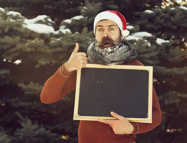 Verrast man in santa hoed, hipster met baard en snor bedekt met witte vorst, met blanco zwart bord of krijtbord geeft duimen omhoog gebaar op winterdag op natuurlijke achtergrond, kopie spa — Stockfoto