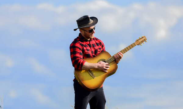 Hiking song. Handsome man with guitar. Country style. Summer vacation. Play beautiful melody. Country music concept. Guitarist country singer stand in field sky background. Inspired country musician