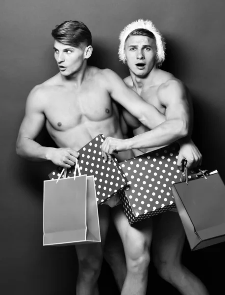 Junge hübsche Macho-Weihnachtsmann überraschte Zwillinge mit sexy muskulös athletisch starken Körper hat nackten Oberkörper und starken Bauch in Weihnachts- oder Weihnachtsmütze mit bunten Geschenktüten auf grünem Studiohintergrund — Stockfoto