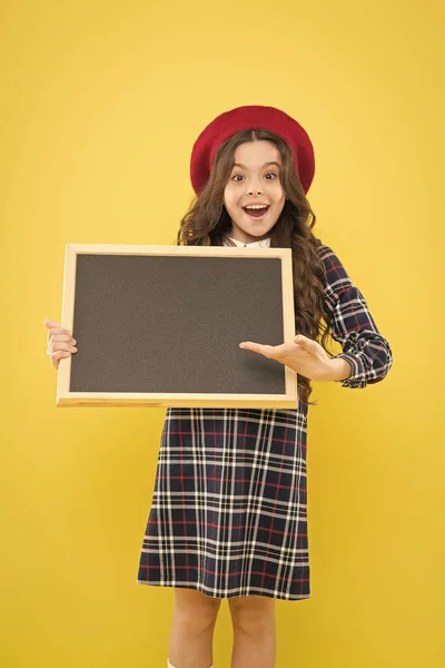 Apresentando o produto. menina pequena em boina francesa. menina feliz com cabelo encaracolado na boina. propaganda. criança com quadro-negro vazio. criança em amarelo. espaço de cópia. Hora da escola. apresentação de projecto escolar — Fotografia de Stock