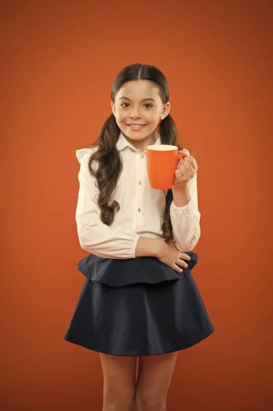 O melhor para ti todas as manhãs. Menina pequena segurando copo com bebida da manhã no fundo laranja. Um miúdo a beber chá matinal ou leite. Criança da escola tomando café da manhã — Fotografia de Stock