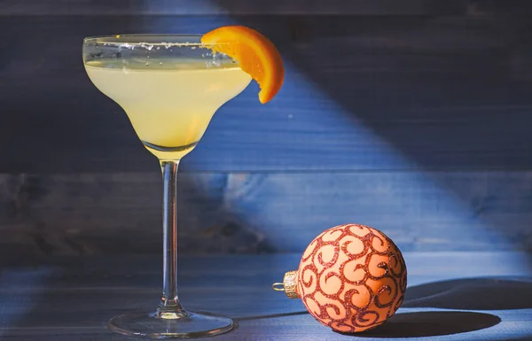 Bar winter season cocktail menu. Winter cocktail drink concept. Alcohol cocktail margarita with piece of orange near christmas ball ornament on dark blue background. Celebrate new year cocktail
