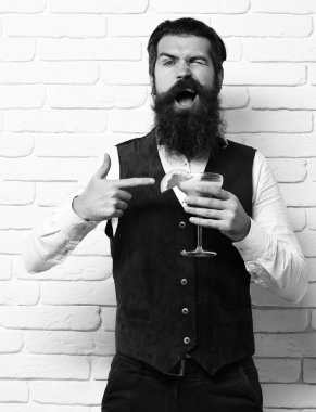 Uzun sakallı ve bıyıklı yakışıklı bir adam komik suratında şık saçlar beyaz tuğlalı stüdyo gerisinde klasik süet deri bir yelek içinde alkollü içecek bardağı tutuyor.