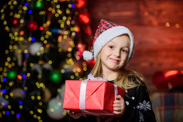Een schattig kind draagt kerstmuts om kerst te vieren. Koop kerstcadeau. Winter winkelen. Meisje schattig kind houden verpakt cadeau in de buurt van kerstboom. Feestje dat eigenlijk leuk klinkt. Vrolijk kerstfeest. — Stockfoto