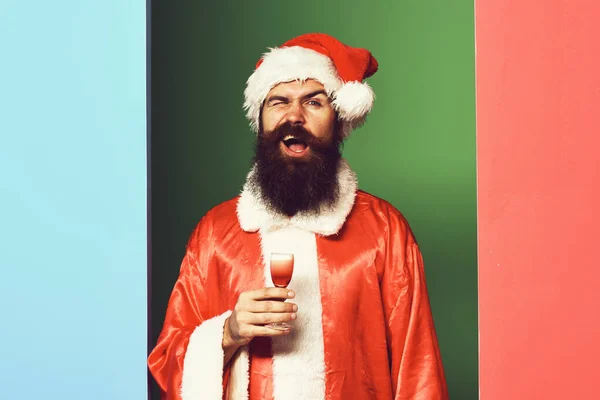 Beau barbu santa claus homme avec longue barbe sur le visage drôle tenant verre de coup alcoolique en rouge Noël ou chandail de Noël et chapeau de nouvelle année sur backgrou studio coloré — Photo