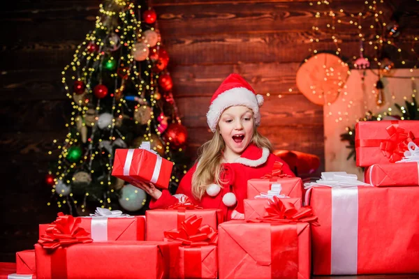 Дитяча щаслива збуджена дівчина знаходить подарунки біля ялинки. Щастя і радість. Веселого Різдва. Щаслива концепція дитинства. Дитячий одяг Санта-капелюх тримає загорнуту різдвяну подарункову коробку. Санта приніс мені подарунки — стокове фото