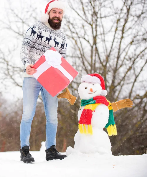 Guy happy face snowy nature background. Hipster with beard hold gift box. Man made snowman. Man Santa hat having fun outdoors. Surprise concept. Winter games. Winter activity. Winter vacation — Stock Photo, Image