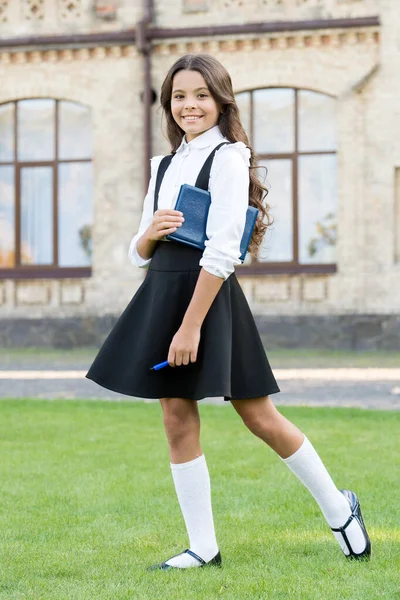 Lección aprendida. Feliz niñita de vuelta a la escuela. Pequeño niño mantenga libro y pluma al aire libre. Linda sonrisa de niño pequeño a la hora de la escuela. Adorable colegiala pequeña con pelo largo morena en ropa formal — Foto de Stock