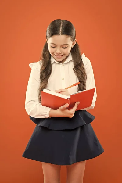 Školačka píše poznámky na oranžové pozadí. získat informační knihu. Zpátky do školy. šťastné dítě se soustředilo na práci. lekce čtení. malá holka ve školní uniformě. Taková zajímavá kniha. — Stock fotografie