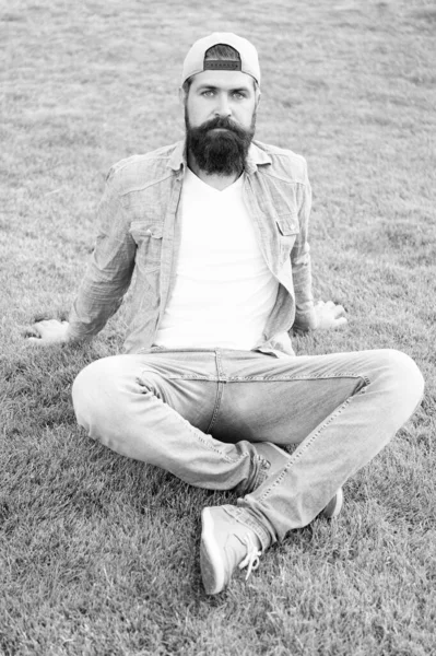 Hårig hipster. Brutal hipster sitter på grönt gräs. Bearded man i trendig hipster stil på sommaren utomhus. Kaukasisk hipster med tjockt skägg hår klädd i keps — Stockfoto