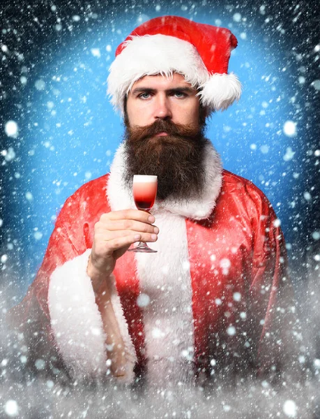 Όμορφος γενειοφόρος Άγιος Βασίλης άντρας με μακριά γενειάδα σε σοβαρό πρόσωπο κρατώντας ένα ποτήρι αλκοολούχο σφηνάκι σε κόκκινο χριστουγεννιάτικο πουλόβερ ή χριστουγεννιάτικο πουλόβερ και καπέλο του νέου έτους σε μπλε φόντο στούντιο — Φωτογραφία Αρχείου
