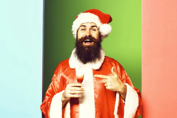 Bel homme barbu santa claus avec une longue barbe sur le visage souriant tenant verre de coup alcoolique en rouge Noël ou manteau de Noël et chapeau de nouvelle année sur backgro studio coloré — Photo