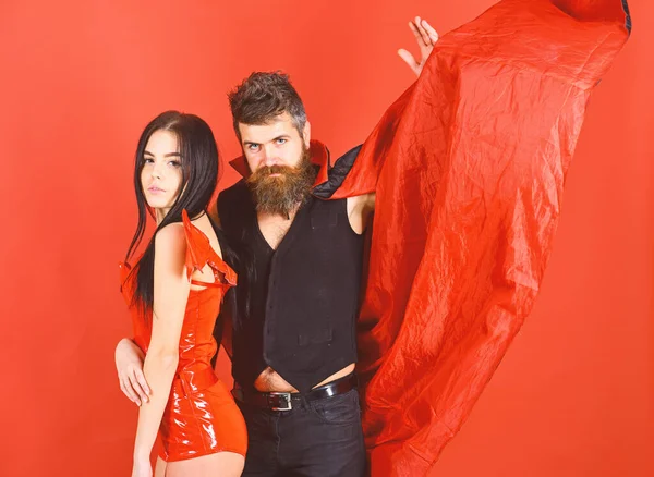 Attractive demon concept. Couple on strict face play role game. Vampire in cloak and sexy devil girl cuddling. Couple in love, perfect match. Man and woman dressed like vampire, demon, red backgroun