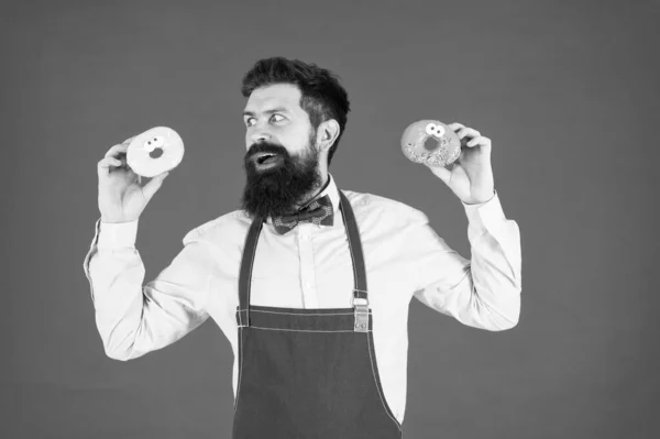 Un beignet glacé. Homme barbu bien entretenu dans un tablier vendant des beignets. Nourriture aux beignets. Des pâtisseries. Bonbons et gâteaux. De la malbouffe. Hipster barbu boulanger tenir des beignets. Doux beignet. Serveur dans un café. Calories de beignets — Photo