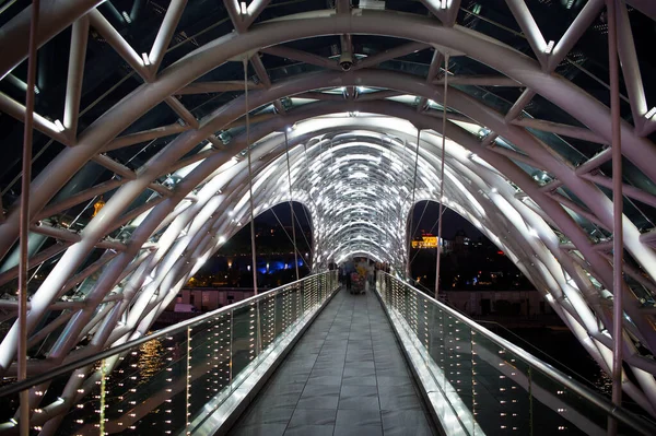 Πεζόδρομος γέφυρα. Χαλύβδινες κατασκευές φώτιζαν πολλά φώτα. Αρχιτεκτονική έννοια. Γέφυρα στην Τιφλίδα. Καμπυλωτό γυάλινο θόλο λαμπυρίζει το βράδυ. Σύγχρονη σχεδιαστική γέφυρα. Ταξιδιωτικοί προορισμοί — Φωτογραφία Αρχείου