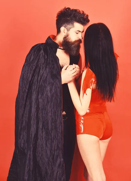 Vampire in cloak and sexy devil girl holds hands. Couple in love, perfect match. Couple on pensive faces play role game. Man and woman dressed like vampire, demon, red background. Halloween concep