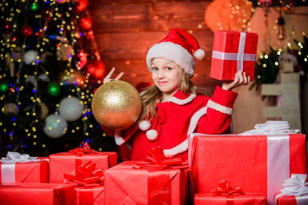 Дитячий одяг Санта-капелюх тримає загорнуту різдвяну подарункову коробку. Санта приніс мені подарунки. Щастя і радість. Дитяча щаслива збуджена дівчина знаходить подарунки біля ялинки. Веселого Різдва. Концепція щасливого дитинства — стокове фото