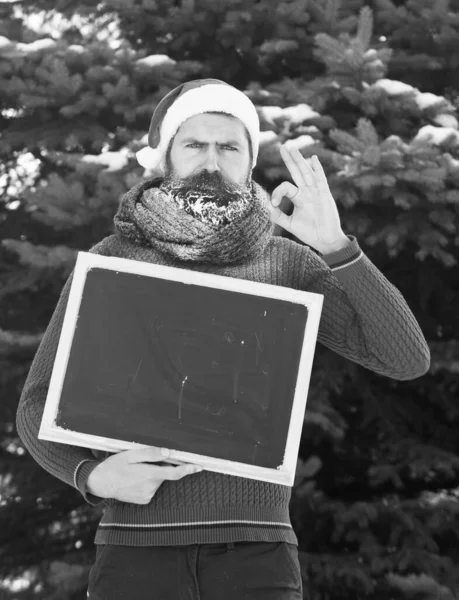 Ernster Mann mit Weihnachtsmannmütze, Hipster mit Bart und Schnurrbart mit weißem Reif bedeckt, mit schwarzer Tafel oder Kreidetafel gibt an einem Wintertag auf natürlichem Hintergrund OK-Geste, Thermalbad kopieren — Stockfoto