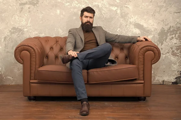 Rent apartment. Bearded man with confident face sit leather couch. Loft interior apartment. Businessman realtor work. Furniture shop. Hipster realtor loft style apartment. Realtor and rental service