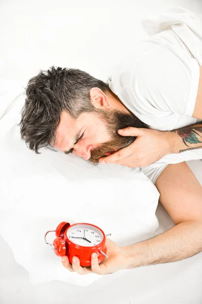 Man with yawning face lies on pillow near alarm clock.