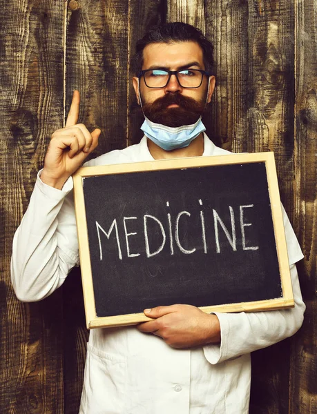 Barbudo brutal caucasiano médico segurando bordo com medicina inscrição no marrom madeira vintage estúdio fundo — Fotografia de Stock