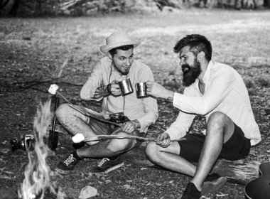 Geleneksel kamp ikramı. Arkadaşlar ormanda şenlik ateşinin yanında dinleniyorlar. Erkekler çubuklarda marşmelov kızartıyorlar. Marshmallow popüler tatlı. Geleneksel kamp işi şenlik ateşinde marşmelov kızartıyor.