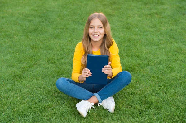 Yeni kitaptan ilham alan genç bir kız. Yeşil çimlerin üzerinde rahatla. Bahar tatili zamanı. Mutlu çocukluk. Çocuk dışarıda parkta kitap okuyor. Küçük mutlu kız tatilde ders çalışıyor. çocuk güzelliği ve moda — Stok fotoğraf